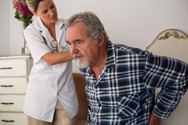Female doctor assisting senior patient experiencing back pain in a retirement home. Ideal for use in healthcare, elderly care, medical assistance, and home care contexts. Can be used in articles, brochures, and websites focused on senior health, caregiving, and medical services.