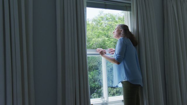 Senior woman stands by window in home, looking outside. Suitable for themes of aging, solitude, mindfulness, and peaceful home environments.