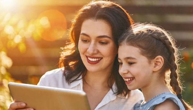 Mother and daughter sharing tablet in sunlit backyard during golden hour