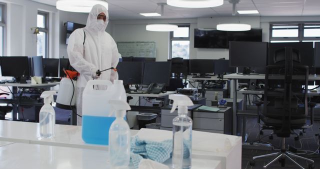 Worker dressed in full protective gear, disinfecting a modern office space. Fingering empty desks, while various cleaning supplies are seen in the foreground. Suitable for use in articles or advertisements about workplace hygiene, safety, pandemic response, healthcare protocols, or office maintenance.