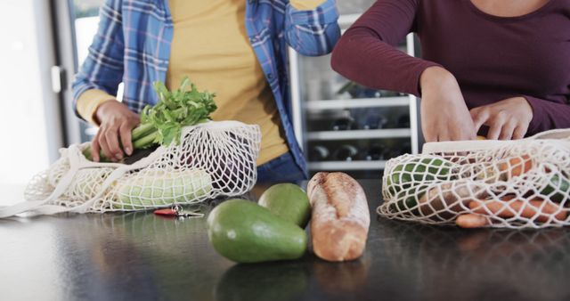 People unpacking groceries from reusable mesh bags at home, emphasizing eco-friendly and sustainable living. Fresh vegetables, avocados, and loaf of bread are present on the table. Suitable for topics related to environmentally conscious shopping, healthy eating, and modern lifestyle. Perfect for magazines, blogs, or marketing materials promoting sustainability and health.