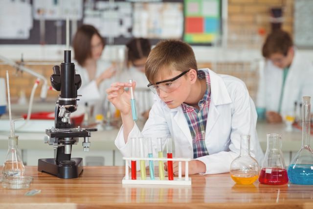 Schoolboy wearing safety goggles and lab coat, carefully examining test tube in school laboratory. Background shows classmates engaged in similar activities. Ideal for educational content, science-related articles, and promotional materials for STEM programs.