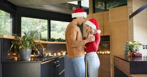 Couple wearing santa hats dancing and holding hands in modern kitchen with festive lights