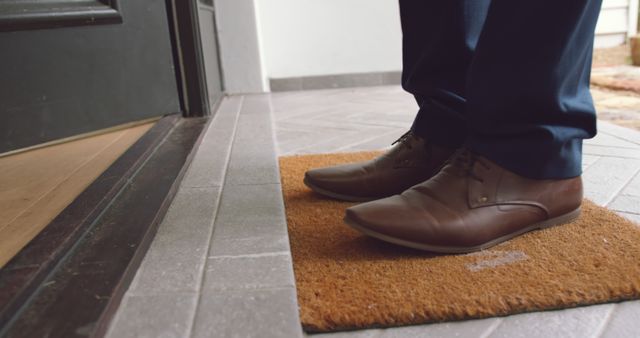 Close-up of a person's feet wearing brown leather shoes standing on a doormat by an entrance door. Ideal for use in advertisements for footwear, business attire, or indoor furnishings. Great for illustrating concepts of arrival, hospitality, or professional settings.