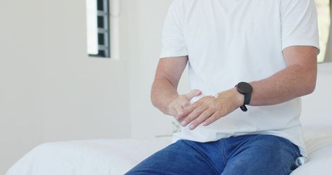 Mature man applying hand lotion while sitting on bed in bright minimalist bedroom interior