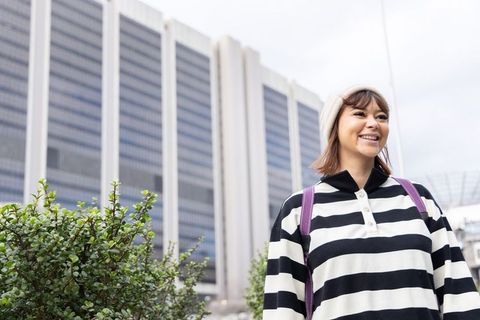 Young woman smiling in urban plaza wearing beanie and striped sweater with backpack