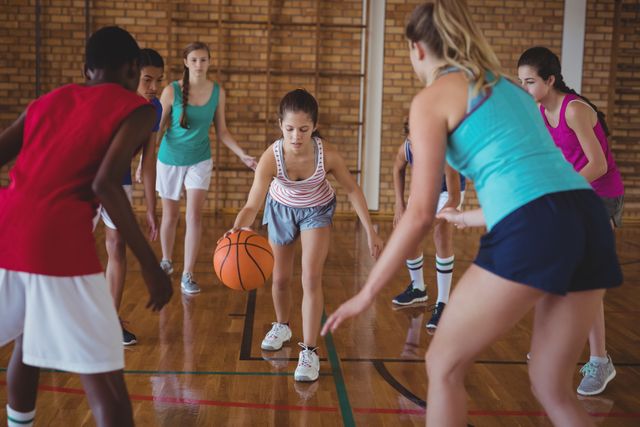 Group of high school students actively playing basketball on an indoor court. Youth athletes engaged in dribbling and teamwork during a game. Perfect for topics related to physical education, youth sports, teamwork, school activities, and healthy lifestyles.