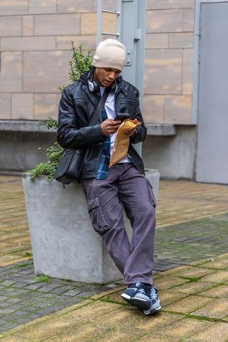 Urban man leaning on planter checking smartphone holding paper bag wearing leather jacket
