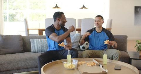 Diverse friends enjoying game day pizza and fist bump on couch in modern living room