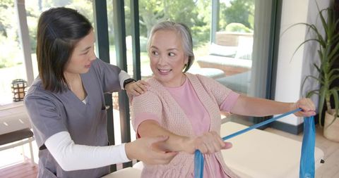 Senior Asian woman exercising with resistance band while caregiver guiding rehabilitation