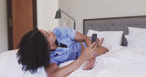 African woman relaxing on bed in blue satin pajamas scrolling smartphone in modern bedroom