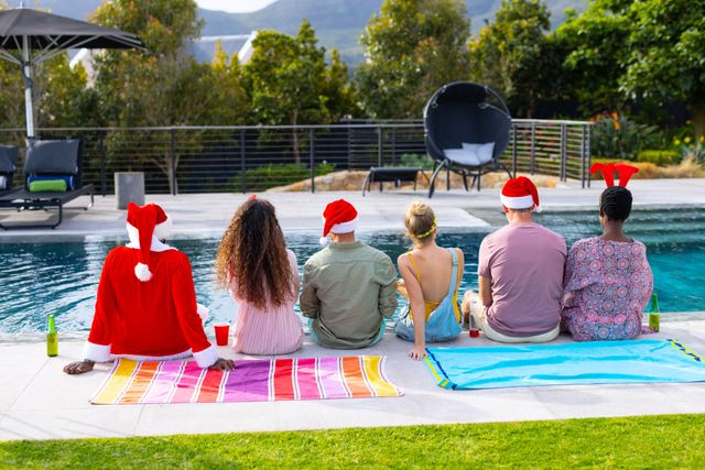 Rear view of diverse group of friends in chirstmas hats sitting by pool with drinks, copy space. Christmas, celebration, friendship and togetherness concept.