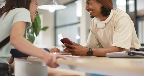 Friends smiling and sharing smartphone at cafe counter with headphones and notebooks