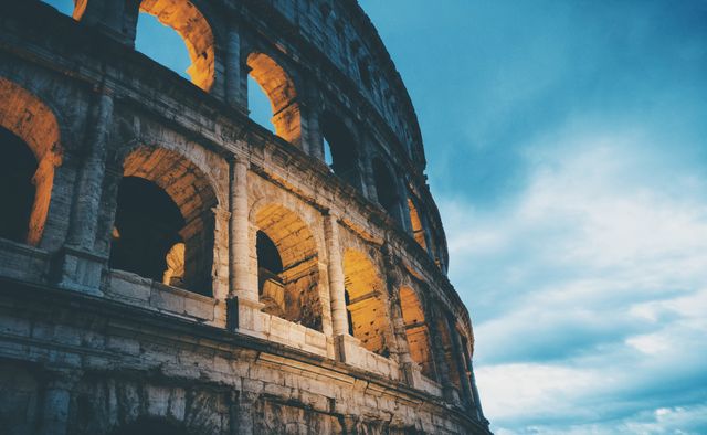 Colosseum's illuminated arches stand against dramatic evening sky. Use in travel blogs, history features, educational content on Roman architecture, promotion of cultural heritage, or as a visual reference for iconic landmarks.