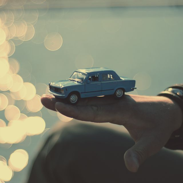 A person is holding a vintage blue toy car in their hand. The background features sparkling sunlight reflecting on water with a bokeh effect, creating a pleasant outdoor scene. Ideal for use in themes about nostalgia, childhood memories, leisure activities, or summer fun.