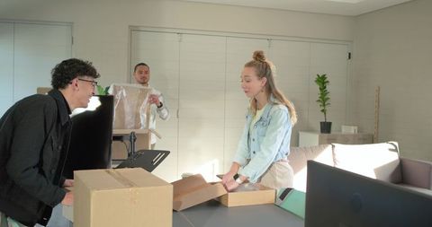 Roommates unpacking new monitor in sunlit modern apartment living room, teamwork unboxing
