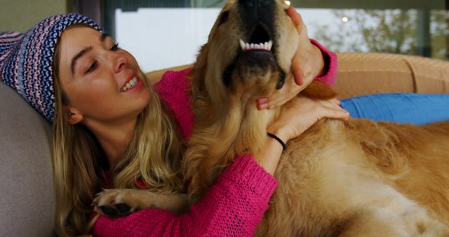 Young woman enjoying quality time and playing with her golden retriever dog on a couch indoors. She wears a knitted beanie and pink sweater, creating a cozy and casual atmosphere. This image can be used for illustrating topics related to pet love, companionship, indoor relaxation, and lifestyle bonding moments with pets.