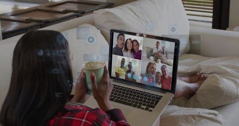 Woman relaxing on sofa drinking coffee while joining family group video call on laptop