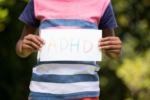 Child holding a sign with ADHD written in colorful letters, standing in a park. Useful for articles on ADHD awareness, mental health education, and support for children with ADHD. Can be used in educational materials, awareness campaigns, and health-related websites.