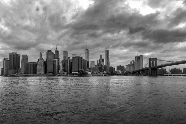 Black and white image of New York City's skyline featuring the Brooklyn Bridge. Tall skyscrapers of Manhattan stand behind the bridge under a dramatic, cloudy sky. This image is ideal for projects needing dramatic urban scenes, cityscape backgrounds, travel articles, or historic perspectives of New York landmarks.