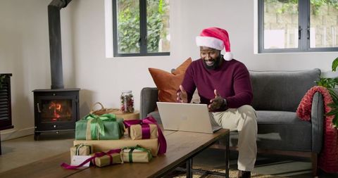 African American man wearing Santa hat video calling family on laptop in cozy living room