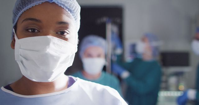 Confident female surgeon wearing face mask in operating room. Dressed in surgical attire, she represents professional dedication. Other medical professionals blurred in background, highlighting teamwork. Ideal for healthcare publications, medical websites, and educational materials related to surgery and hospitals.