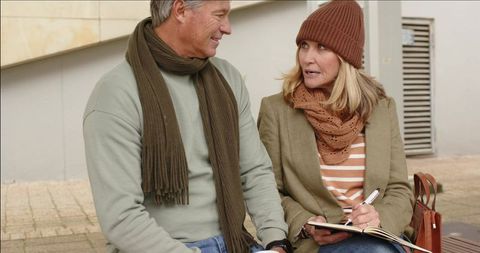 Senior couple journaling and chatting on urban bench wearing scarves and taking notes