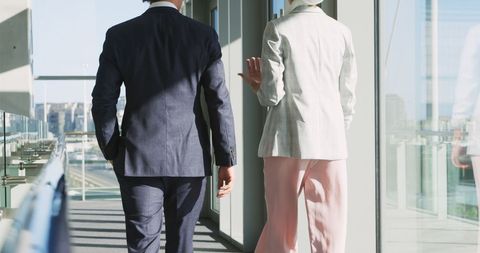 businessman and businesswoman are actively engaging in a work-related discussion while walking through an office corridor. dressed in formal attire, their animated conversation highlights teamwork and collaboration in a modern office setting. the cityscape visible through the glass windows adds to the contemporary business environment. useful for portraying professional communication, teamwork, and corporate settings.