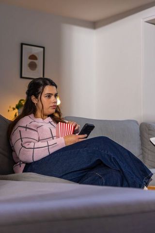 Young woman relaxing on sofa with popcorn and smartphone during cozy evening streaming