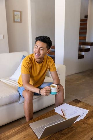 Young asian man in mustard t-shirt sitting on sofa holding mug near laptop smiling