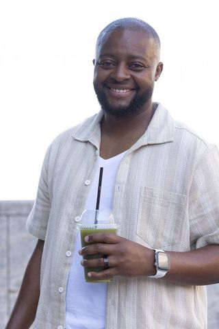 Smiling man holding green juice in takeaway cup wearing casual shirt and smartwatch on rooftop