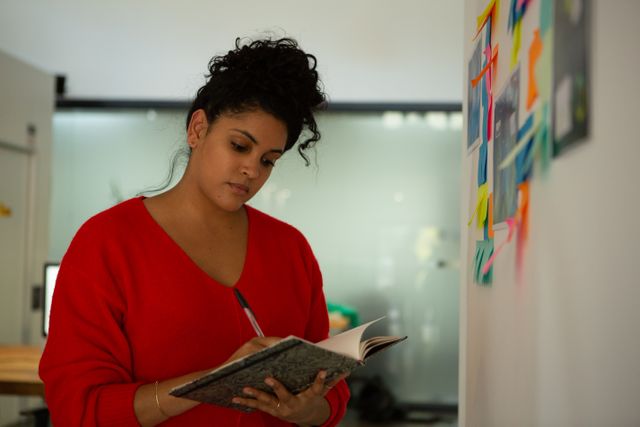 Side view of a biracial woman working in a creative office, standing by a wall with memo cards and posters on it, writing things down in a notebook