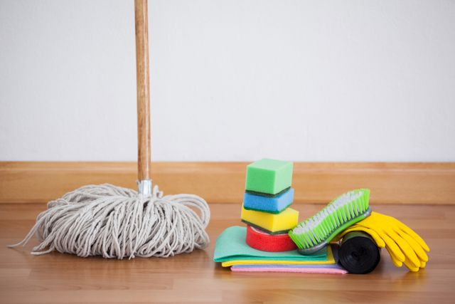 Various cleaning supplies including a mop, sponges, gloves, and a cleaning brush are arranged on a wooden floor. Ideal for illustrating household chores, cleanliness, and hygiene. Suitable for use in articles, advertisements, and blogs related to home cleaning and maintenance.