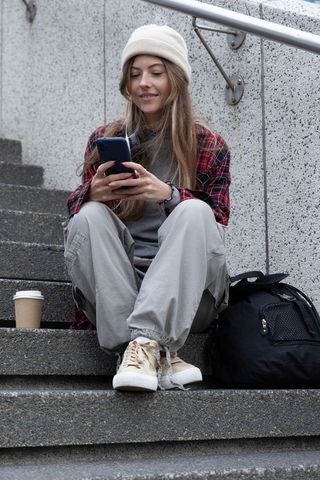 Young woman in beanie sitting on concrete steps scrolling smartphone with backpack and coffee