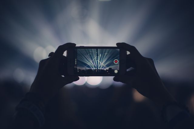 Person recording concert with a smartphone while dramatic light beams shoot from the stage in background. Audience and live performance atmosphere makes it great for creativity, technology in everyday life, or social media themed material. Use in blogs, promotional materials for concerts, or technology related content.