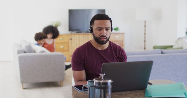 This photo depicts a man working from home, sitting at a table with a laptop and wearing a headset for a virtual meeting. A French press, phone, and documents are also on the table. In the background, a woman and a child can be seen on a couch, suggesting a family environment. Use this for articles or advertisements focused on remote work, work-life balance, or modern family dynamics in the digital age.