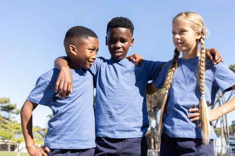 Diverse school friends linking arms on sunny field in blue t-shirts and shorts