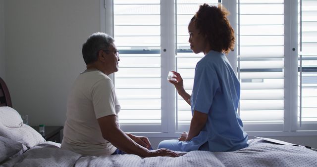 Nurse is giving medicine to a senior man sitting on bed in a bedroom with natural light coming through windows. Ideal for topics related to elderly care, healthcare services, home nursing, and caregiving support.