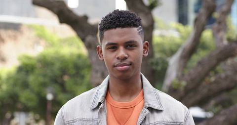Confident african american teen standing in city park wearing denim jacket and orange sweater