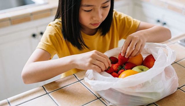 Schoolgirl selecting strawberries from produce bag on tiled kitchen counter for healthy snack