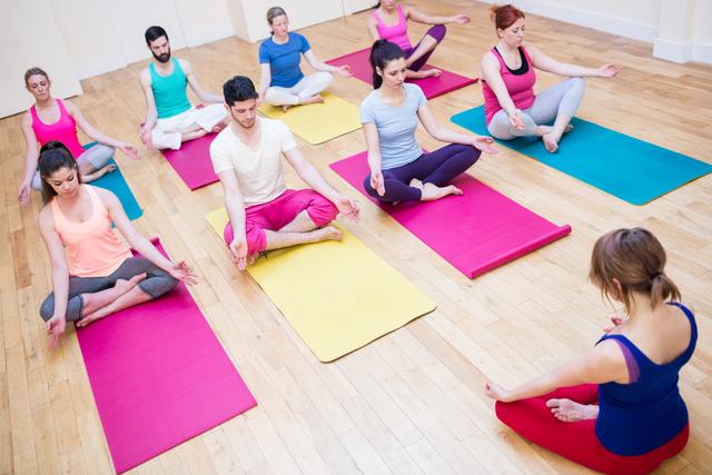 Group of people practicing meditation in a fitness studio, sitting in lotus position on colorful yoga mats. Ideal for promoting wellness programs, yoga classes, mindfulness training, and healthy lifestyle initiatives.