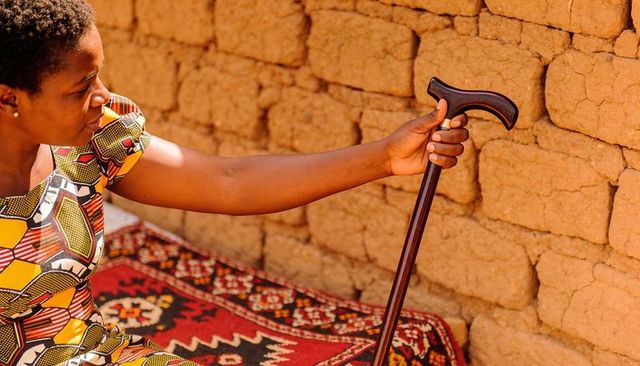 Woman holding wooden cane by adobe wall in sunlit courtyard wearing colorful waxprint dress
