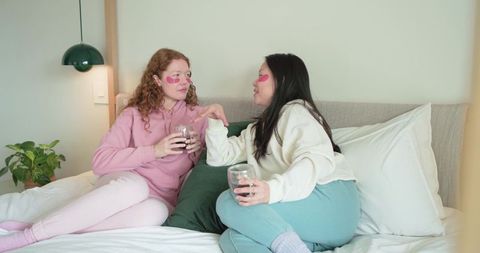 Two friends relaxing on bed with pink under-eye patches sipping wine in pastel bedroom