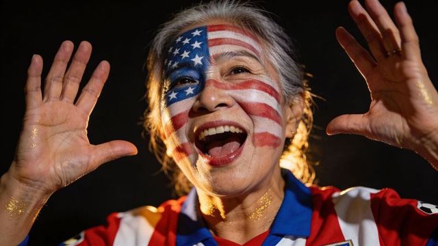 Senior asian fan cheering with american flag face paint and gold glitter at night game