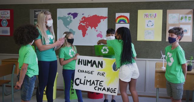 Children in green shirts and masks in a classroom raising awareness for climate change by holding a recycling symbol banner and having group discussion with a teacher. Useful for topics related to climate education, student environmental initiatives, school recycling programs, and youth activism.