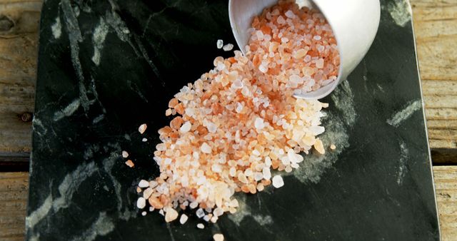Pink Himalayan salt is spilled from a white container onto a dark, textured surface, with copy space. The image captures the natural beauty and color variation of the gourmet salt, often used for culinary and health purposes.