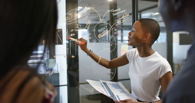 Businesswoman explaining financial graphs to colleagues on transparent board in modern office. Ideal for business presentations, financial strategies, corporate meetings, teamwork, and professional collaboration themes.