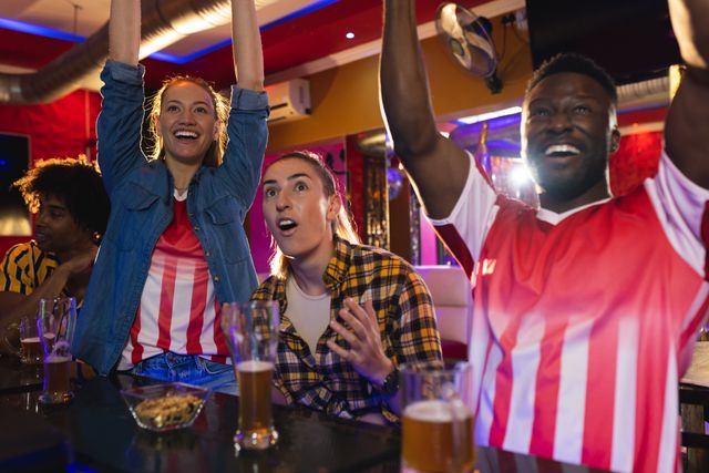 Group of diverse friends enthusiastically cheering while watching a sports game in a lively pub. Ideal for use in advertisements for sports bars, social gatherings, or promoting camaraderie and friendship.