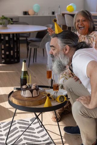 Older couple celebrating birthday at home man blowing candles on chocolate cake