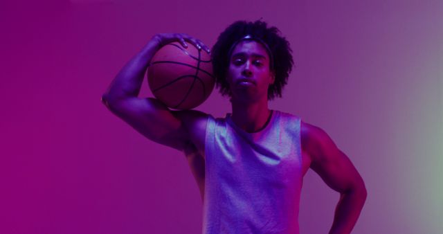 Young male basketball player confidently posing with ball under neon purple lighting. Great for content related to sports, fitness, determination, athleticism, modern fashion, and youth culture advertising.